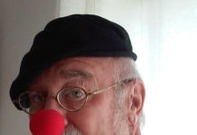 Stuart Goldman is a white man with grey beard. He is wearing an orange shirt, black hat and red clown nose.
