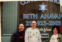 Standing in front of a window with "Congregation Beth Ahavah" painted on it, three white people stand dressed in sweatshirts.