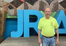 A white man in jeans and a neon yellow short stands in front of large block letters that read "JRA".