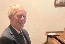 A white man with white hair and glasses smiles and plays a light brown grand piano.