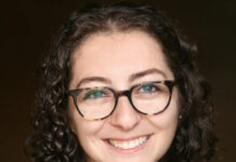 Ariel Milan-Polisar is a white woman with glasses and very curly hair, wearing a black blazer and green blouse.