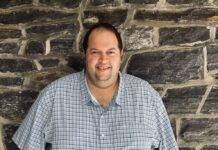 A man in a blue shirt stands in front of a brick wall.