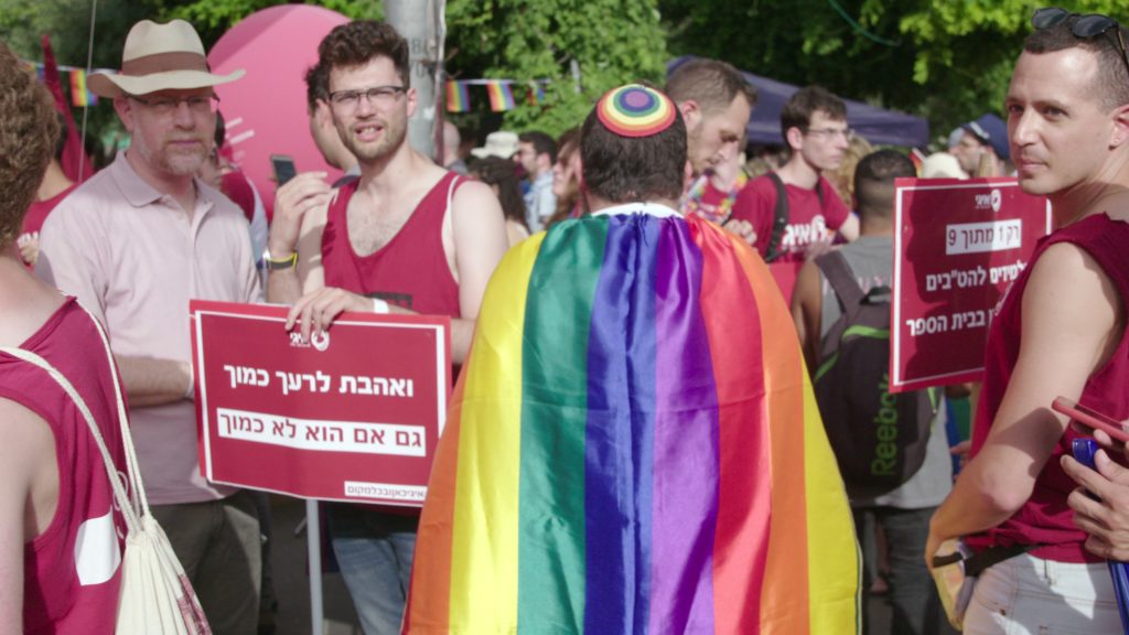 image of man in yarmulke wearing a rainbow flag with other people holding signs in hebrew