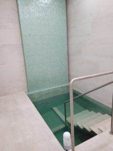 The women's mikvah is a large, square bath with teal green tiling. It is in a larger building with large, marble blocks. There is a marble staircase leading to the mikvah, with two handrails.