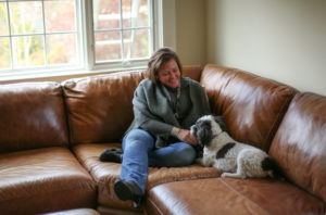 Virginia Buckingham is a white woman with brown hair above her shoulders. She is relining on a light brown leather couch and petting her dog.
