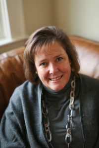 Virginia Buckingham is an older white woman with bronw hair above the shoulders and wispy bangs. She is smiling at the camera and wearing a grey turtleneck and jacket with a piece of chunky jewelry around her neck.