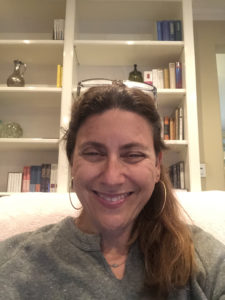 Audrey Kraus is a white woman with long, light-brown hair tied back in a ponytail. She has glasses propped on top of her head and is wearing hoop earrings. She is smiling at the camera.