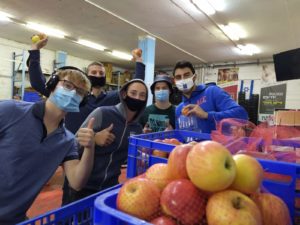 Behind a blue crate filled with apples, several masked students look at a camera, smiling behind masks. One boy in the front is giving a thumbs up.