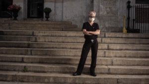 Natanya Gornstein-Talotti is a white woman wearing glasses, a surgical head covering and navy blue scrubs and a mask. She has her arms crossed and is standing on the steps on her school, Penn Alexander.