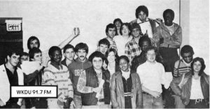 A black and white pictures shows several students from the radio station smiling; some are holding up cans.
