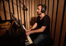 Emiliano Messiez is an Argentine man with a short hair and beard wearing a black t-shirt and has headphones over his ears. He is playing the piano.