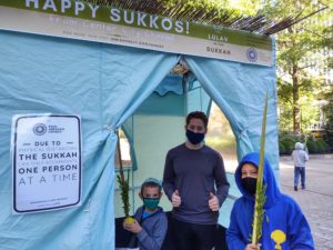 Standing in front of a sukkah draped in blue fabric is a man in a purple shirt and two kids, one holding an etrog, the other holding a lulav; everyone is wearing a face mask.