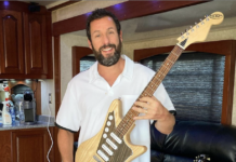 Adam Sandler is a white man with dark hair and a dark beard with a few grey streaks in it. He is wearing a short-sleeved button-up shirt and red shorts and is holding a custom guitar.