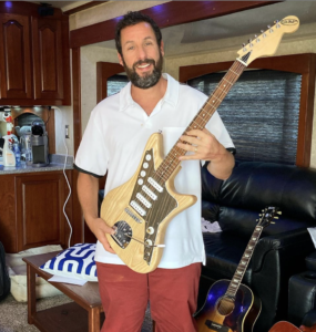Adam Sandler is a white man with dark hair and a dark beard with a few grey streaks in it. He is wearing a short-sleeved button-up shirt and red shorts and is holding a custom guitar.