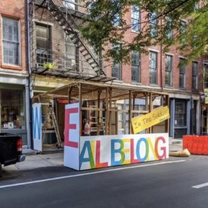 Next to a Philadelphia street, a wooden sukkah stands. It is painted in rainbow letters to say "WE ALL BELONG" across it.