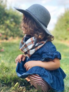 A toddler with shoulder-length, curly hair is crouched on grass outside. She is wearing striped leggings, a denim dress, and a wide-brimmed hat.