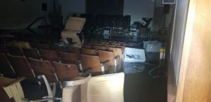 A dark room lit by a flashlight has water rising around wooden chairs in an auditorium. Bins and office chairs are floating in the brackish water.