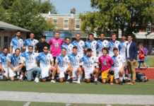A group of over 20 men standing and kneeling in two rows pose for a picutre. They are wearing blue and white soccer jerseys, except for two goalies, who are wearing pink shirts. Coach Tyler Weiss is smiling to the right of the players, wearing a suit.