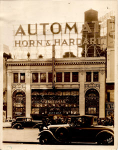 A sepia-toned photograph shows a large building on the streets of New York with black cars rushing by it. At the top of the building, there is a large sign that says, "AUTOMAT HORN & HARDART".