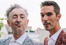 Alan Cumming (left) is a white man with slicked back grey hair sneering playfully at the camera wearing a grey plad suit. Next to him, Ari Shapiro, a younger man with dark, cropped hair is smiling and wearing a red plaid suit.