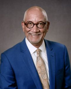Les Robbins is a light-skinned Black man with thick, round classes and grey stubble on his face and head. He is wearing a blue suit and gold tie and is smiling.