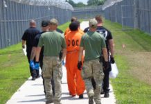 Two men in green and camo lead an incarcerated individual in orange down a long, wide sidewalk surrounded by tall, chainlink fence.