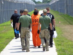 Two men in green and camo lead an incarcerated individual in orange down a long, wide sidewalk surrounded by tall, chainlink fence.