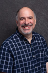 Len Lipkin is a white man with thinning grey hair and a goatee wearing a blue plaid shirt. He is smiling in front of a grey background.