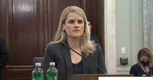 Frances Haugen is a white woman with blend hair wearing a grey blazer. She is sitting in front of a microphone and two plastic water bottles.