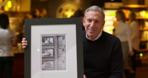 Howard Schultz is a middle-aged white man wearing a white shirt under a blue sweater. He is smiling and holding a framed black-and-white photo of the Automat's food dispensing cubbies.