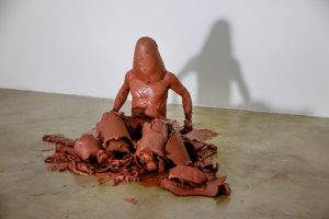 A brown clay sculpture of a crude man sitting and beginning to stand up is photographed in front of a white background