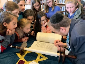 Rabbi Yonah Gross is a white man with light brown hair wearing a suit and editing a Torah at a desk, surrounded by young children looking over him. 