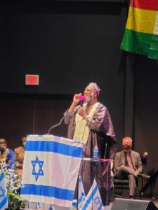 Rev. Dumisani Washington is a black man standing in front of a podium with an Israeli flag onstage . He is speaking animatedly.