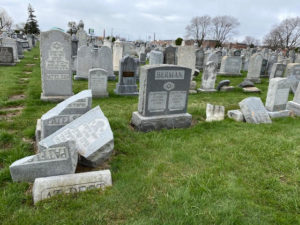 In a cemetery with green grass and small, bare trees, several grey gravestones are standing, but many are toppled or broken into multiple pieces.