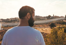 A Palestinian man with short, dark hair and full beard wearing a white t-shirt is standing in front of a large field and border wall.