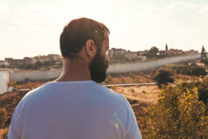A Palestinian man with short, dark hair and full beard wearing a white t-shirt is standing in front of a large field and border wall.