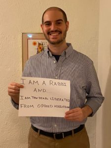 Rabbi Michael Perice is a white man with short, thinning hair and a short beard. He is wearing a button-up shirt and is wearing a sign that reads, "I am a rabbi, and I  am ten years liberated of opioid addiction."
