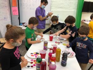 Five children in masks are painting on a table filled with paint bottles and plates. They are supervised by an educator, also wearing a mask.
