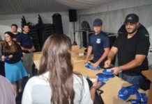 Two men wearing baseball caps and blue shirts and standing behind a wooden table and are handing out blue bags filled with Narcan.
