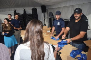 Two men wearing baseball caps and blue shirts and standing behind a wooden table and are handing out blue bags filled with Narcan.