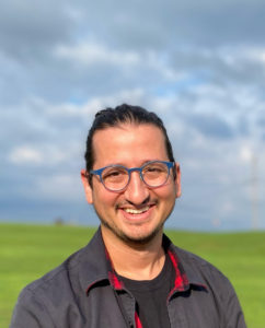 Eli Robbins is a cuban Jew with his dark hair tied back in a bun. He is smiling and wearing blue-rimmed glasses and standing in a field.