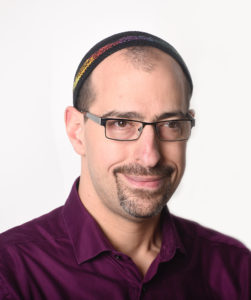 Rabbi Ilan Glazer is a white man wearing a large kippah over short, thinning hair. He wears rectangular glasses and a maroon button-up shirt.