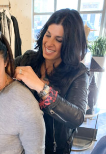 Marlyn Schiff is a white woman with wavy, dark hair wearing a leather jacket and smiling. She is fastening a necklace onto a customer's neck.