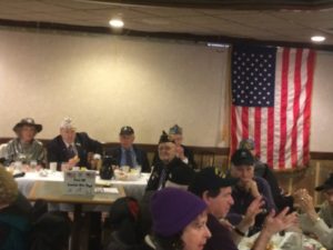 With a large American flag draped behind them, a group of older veterans sit around tables.