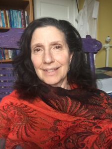 Rabbi Tsurah August is an older white woman with dark eyes, collar-bone length brown hair wearing a red shawl around her shoulders. She is smiling.