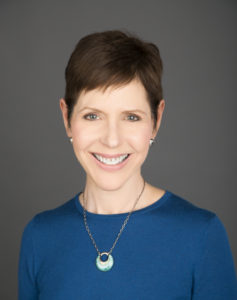 Marla Kaufman is a white woman with short, brown hair, smiling to the camera. She is wearing earrings, a necklace, and a blue shirt.