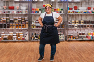 Neomie Eliezer is a Turkish woman with thick, black-rimmed glasses and dark hair tied back in a yellow handkerchief. She is wearing an apron and is smiling with her hands on her hips standing in front of a shelf lined with kitchen supplies.