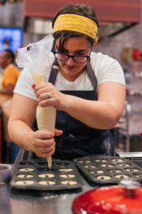 NÃ©omie is holding a pastry bag filled with dough.  She passes it through a circular mold.