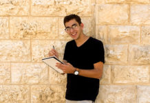 Andrew Galitzer is a white man with short hair and glasses holding a pencil and drawing pad. He is leaning against a stone wall.