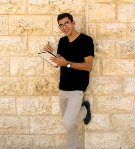 Andrew Galitzer is a white man with short hair and glasses holding a pencil and drawing pad. He is leaning against a stone wall.